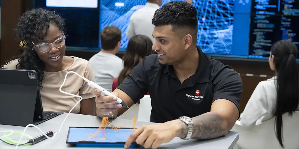 A student builds something with a 3D printing pen, while other watches & references a tablet. A large screen behind them shows a complex network of blue lines.