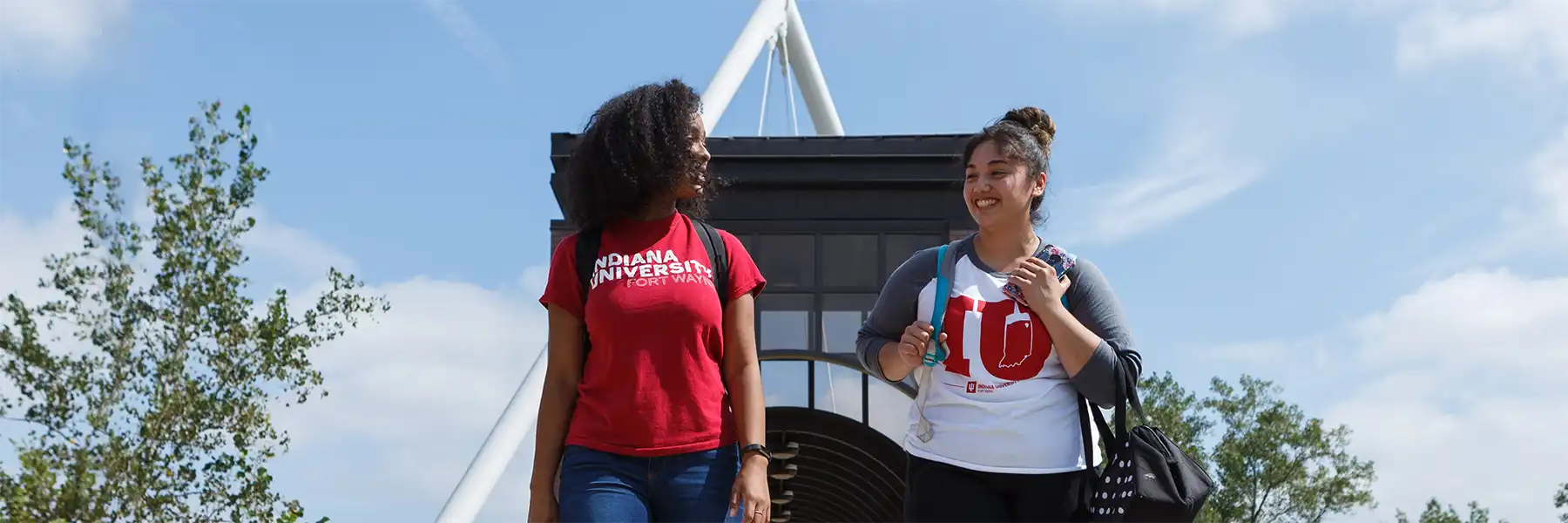 Two IU Fort Wayne students, wearing backpacks and IU shirts, cross a pedestrian bridge while chatting.