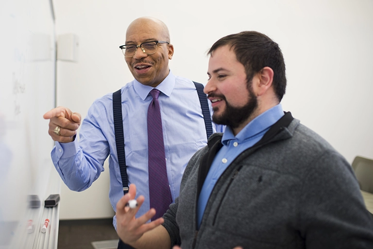 A student and instructor laugh while discussing something written on a whiteboard.