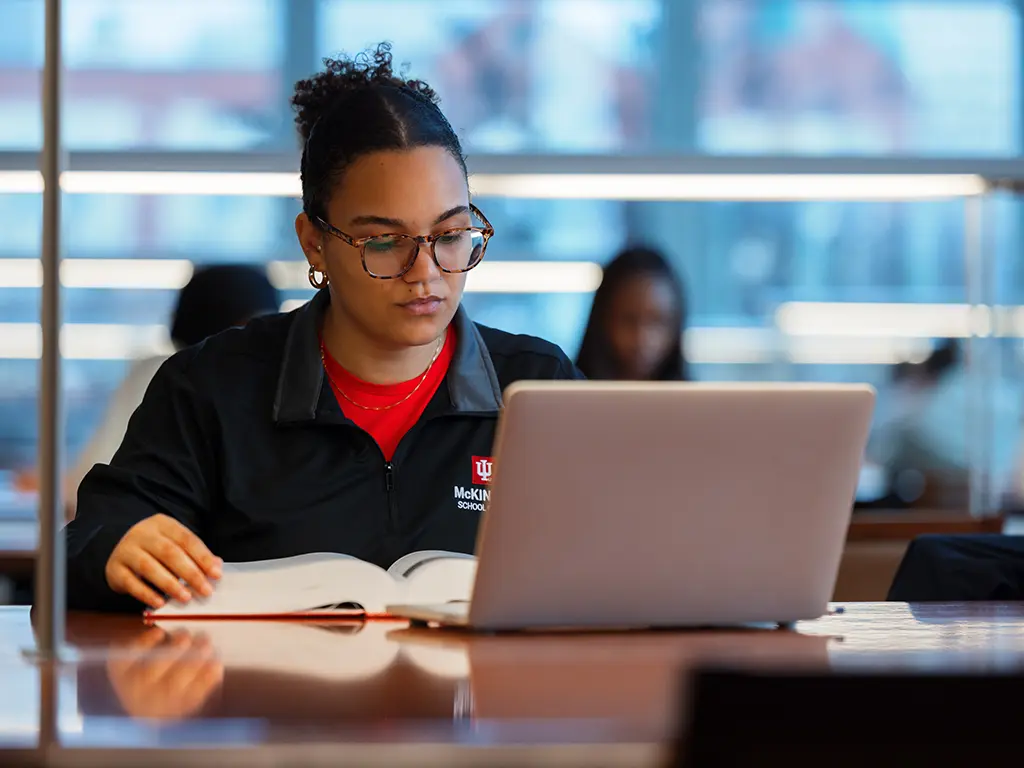 A student reads a thick book in a library. Their laptop is sitting in front of them, open.